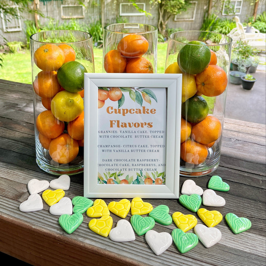 Decorative setup with ceramic hearts scattered in front of glass vases full of citrus fruit and a sign about cupcake flavors.