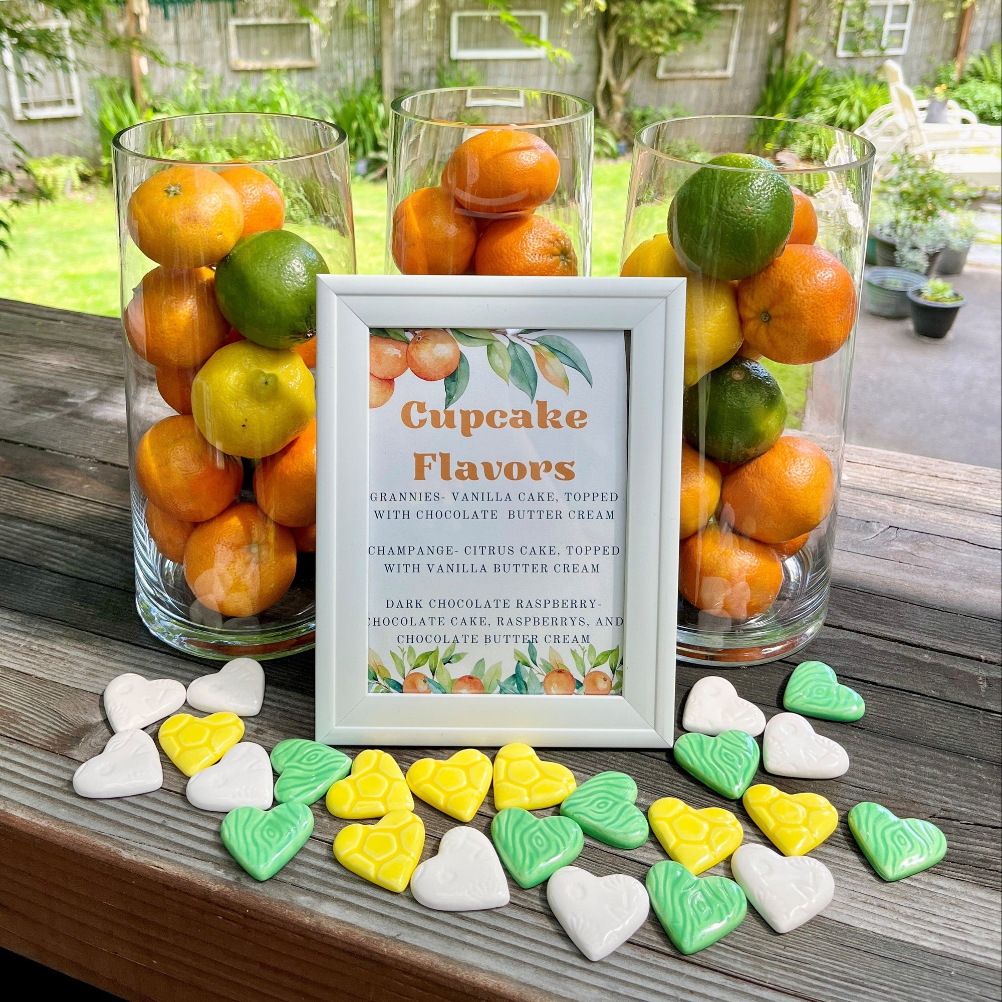 Decorative setup with ceramic hearts scattered in front of glass vases full of citrus fruit and a sign about cupcake flavors.