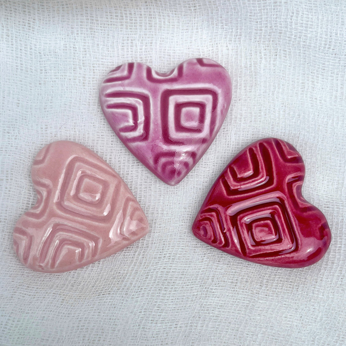 Three ceramic hearts in different pink shades on a white fabric background.