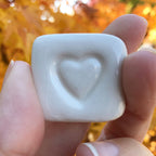 Hand holding a white stone with a heart engraving against an autumn background, labeled 'LOVE STONES'.