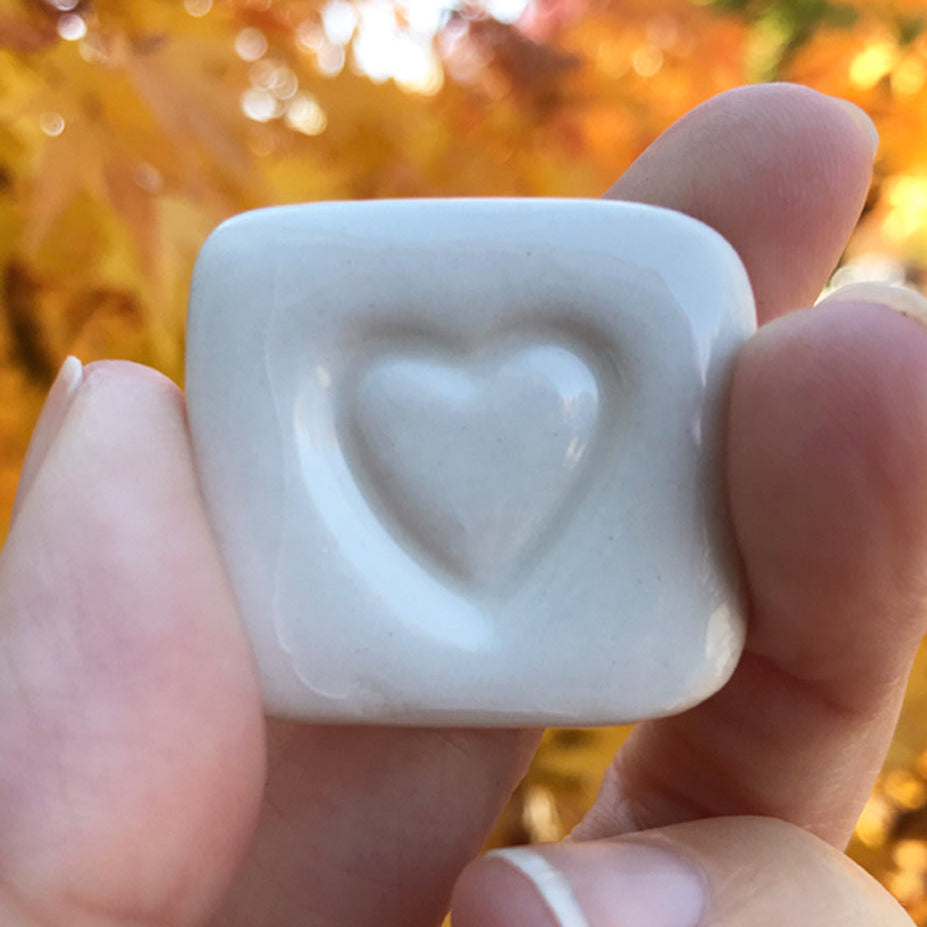 Hand holding a white stone with a heart engraving against an autumn background, labeled 'LOVE STONES'.